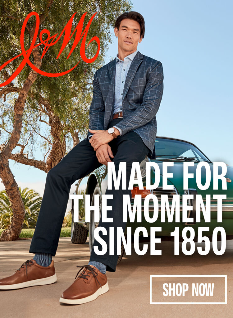Man leaning on a vintage green car wearing a nicer looking outfit with brown shoes, jeans and a blazer. "Made for the moment since 1850" is written on the right side of the banner.